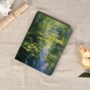 Könnte beinhalten: Ein rechteckiges Notizbuch mit einem impressionistischen Gemälde eines Wassergartens mit Seerosen. Das Kunstwerk zeigt Grüntöne, Blau und Gelb, die ein Gefühl der Ruhe hervorrufen. Das Notizbuch liegt auf einer strukturierten, cremefarbenen Oberfläche.