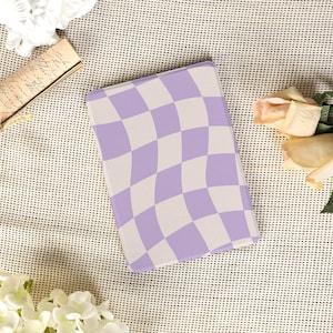 May include: A rectangular notebook with a lavender and white chequered pattern. The notebook has a wavy design and is set against a textured, off-white background. The image also includes flowers and a vintage document.