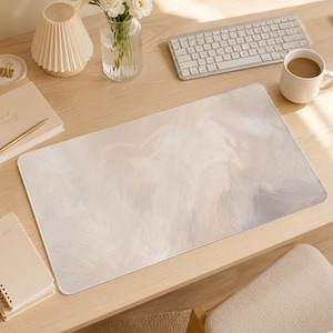 May include: A rectangular desk mat featuring a soft, abstract design with blended strokes of cream, off-white, and light grey. The surface has a smooth, matte texture with rounded corners, providing a neutral and subtle aesthetic for a workspace.