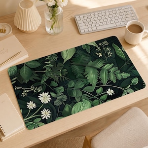 May include: A rectangular desk mat featuring a dark green botanical pattern with various lush leaves, ferns, and small white daisy-like flowers. The design covers the entire surface of the mat, displaying a dense, layered arrangement of foliage.