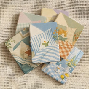 Peut inclure: Une collection de dessous de verre rectangulaires présentant des scènes illustrées d'animaux lisant au lit, un chat avec une couverture à carreaux et une table avec des fleurs jaunes, des citrons et une tasse de thé, dans des tons pastel.