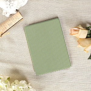 May include: A rectangular notebook with a green and white striped pattern. The notebook is lying on a textured, off-white surface, with flowers and a vintage document nearby. The notebook's cover features vertical stripes.