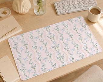 May include: A rectangular desk mat featuring a light pink background with a repeating pattern of vertical wavy lines, small blue floral sprigs, and light blue bows. The mat has rounded corners and a smooth surface finish for desk organization.