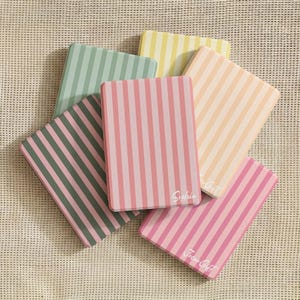 Può includere: Una collezione di quaderni con strisce verticali in vari colori pastello, tra cui rosa, verde, giallo e pesca. Un quaderno ha il nome "Sophia" stampato sopra, e un altro "Jocu Gift". I quaderni sono disposti su una superficie strutturata.