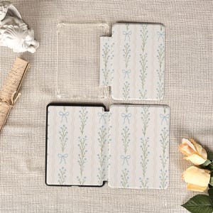 May include: A set of phone cases and a clear phone cover. The cases feature a white background with a repeating pattern of light blue bows and floral stems. The clear cover is visible in the image.