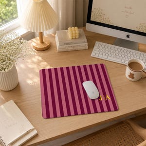 May include: A rectangular mouse pad featuring a pattern of alternating vertical stripes in shades of dark maroon and light pink. The bottom right corner displays the gold-colored initials J and A separated by a vertical line.