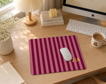 Puede incluir: Una alfombrilla de ratón rectangular con un patrón de rayas verticales alternas en tonos granate oscuro y rosa claro. La esquina inferior derecha muestra las iniciales J y A en color dorado, separadas por una línea vertical.