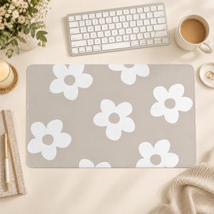 Puede incluir: Una alfombrilla de escritorio rectangular con un fondo beige claro y un patr&oacute;n repetitivo de grandes flores blancas de cinco p&eacute;talos. La alfombrilla tiene una superficie lisa y plana con esquinas redondeadas, ofreciendo un dise&ntilde;o sencillo.
