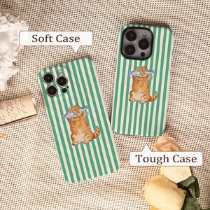 May include: Two phone cases with a green and white striped design. One case is labelled "Soft Case" and the other "Tough Case". Each case features an orange cartoon cat holding two fish. The cases are on a textured surface.