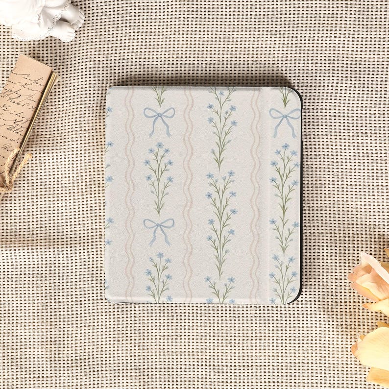 May include: A floral-patterned tablet case featuring a design of light blue flowers and bows on a cream background. The case has a black border and a vertical striped pattern. The overall aesthetic is delicate and feminine.
