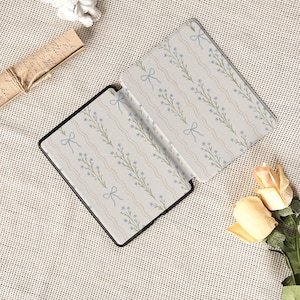 May include: An open black e-reader case with a floral pattern of blue flowers, green stems, and light blue bows on a cream background. The case is open, revealing the interior design. The case is on a textured surface.