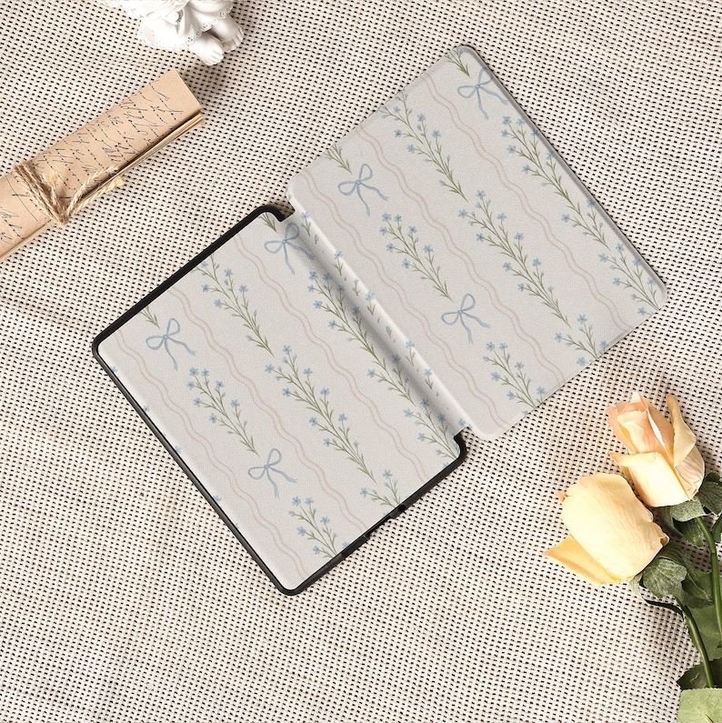May include: An open black e-reader case with a floral pattern of blue flowers, green stems, and light blue bows on a cream background. The case is open, revealing the interior design. The case is on a textured surface.
