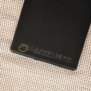May include: Black electronic device with regulatory information printed on the back. The device is rectangular with rounded corners. The surface has a matte finish. The serial number is highlighted with a yellow circle.