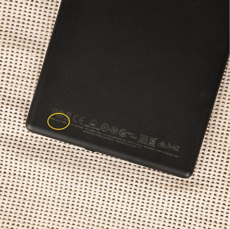 May include: Black electronic device with regulatory information printed on the back. The device is rectangular with rounded corners. The surface has a matte finish. The serial number is highlighted with a yellow circle.