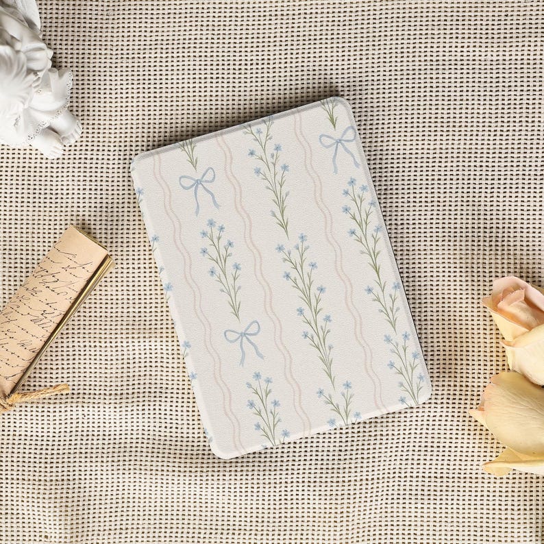May include: A rectangular, off-white decorative item featuring a floral pattern with blue flowers, green stems, and light blue bows. The design includes wavy, light beige stripes. The item is likely a decorative tray or placemat.