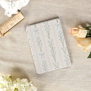 May include: A rectangular, floral-patterned item featuring a repeating design of blue flowers, green stems, and light blue bows on a white background with wavy, light beige stripes. The item is likely a decorative piece or accessory.