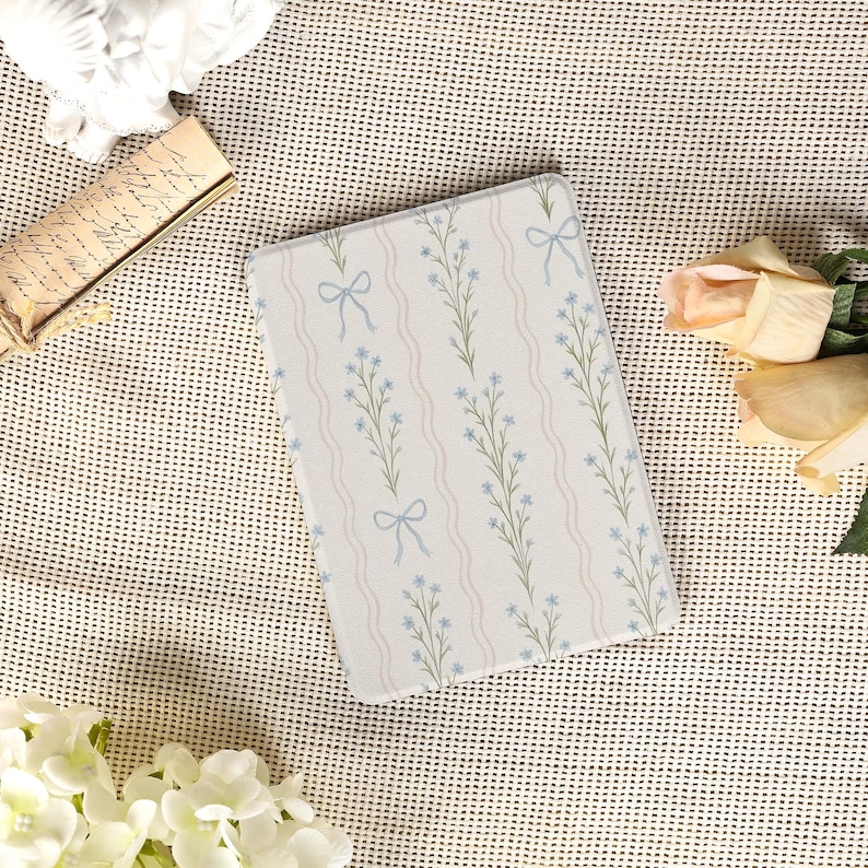 May include: A rectangular, floral-patterned item featuring a repeating design of blue flowers, green stems, and light blue bows on a white background with wavy, light beige stripes. The item is likely a decorative piece or accessory.