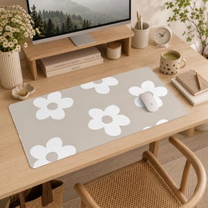 Puede incluir: Una alfombrilla de escritorio rectangular con fondo beige claro y un patr&oacute;n repetitivo de grandes flores blancas de cinco p&eacute;talos con centros beige. La alfombrilla tiene esquinas redondeadas y est&aacute; sobre una mesa de madera con un rat&oacute;n.