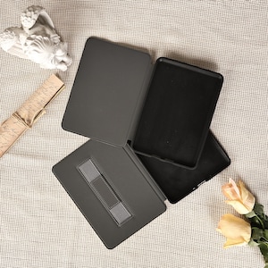 May include: A dark gray tablet case with a built-in hand strap. The case is open, revealing the interior and a black textured surface. The case is designed to protect the tablet from scratches and damage.