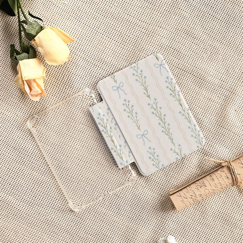 May include: A clear phone case and two notebooks with a floral pattern of blue flowers and bows on a white background. The notebooks have a wavy striped design. The image also includes two yellow roses and a scroll.
