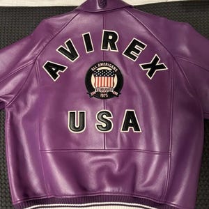 Könnte beinhalten: Eine lila Lederjacke mit dem Wort "AVIREX" über den Schultern. Darunter befindet sich ein Aufnäher mit den Worten "ALL AMERICANS ESTABLISHED 1975 USA". Die Jacke hat einen gerippten Kragen und Bündchen mit weißen Streifen.