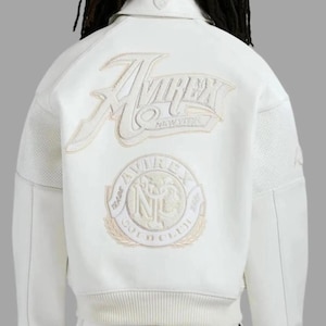 Op de afbeelding: Witte leren jas met het Avirex-logo in crème op de rug geborduurd. De jas heeft een geribbelde zoom en manchetten en een kraag. Het Avirex-logo staat ook op de voorkant van de jas.