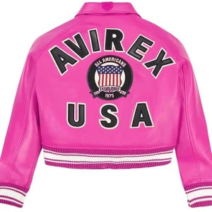Puede incluir: Chaqueta de cuero rosa brillante con la palabra "AVIREX" en letras negras y blancas en la espalda. Debajo, se muestra la palabra "USA". Un parche circular con la bandera estadounidense y las palabras "ALL AMERICANS ESTABLISHED 1975" est&aacute; centrado.