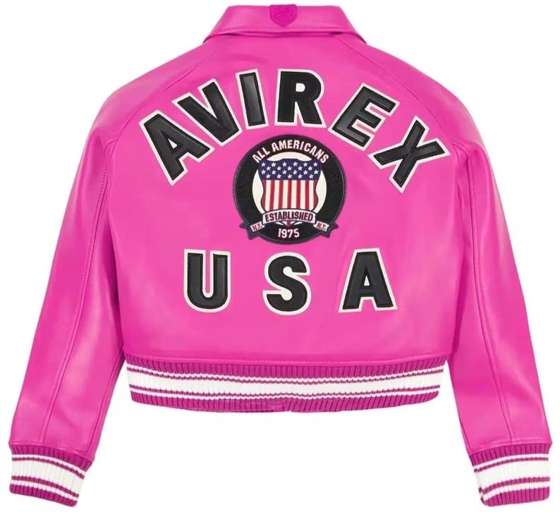 Puede incluir: Chaqueta de cuero rosa brillante con la palabra "AVIREX" en letras negras y blancas en la espalda. Debajo, se muestra la palabra "USA". Un parche circular con la bandera estadounidense y las palabras "ALL AMERICANS ESTABLISHED 1975" est&aacute; centrado.