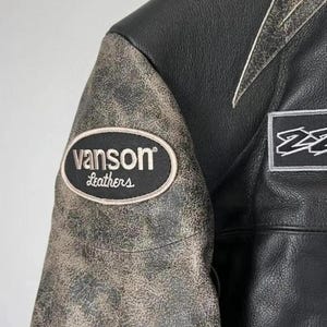 May include: Close-up of a black leather jacket with a distressed, textured sleeve. The jacket features a "vanson Leathers" patch and white accents. The jacket is a classic style, suitable for motorcycle enthusiasts.