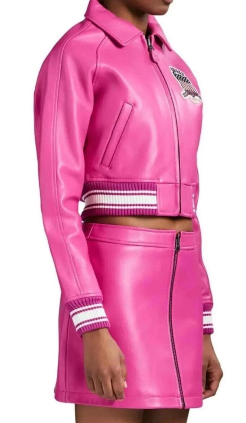 Puede incluir: Conjunto de chaqueta y falda de cuero rosa brillante. La chaqueta tiene cuello, cierre de cremallera y un parche en el pecho. La chaqueta y la falda tienen pu&ntilde;os y cinturillas acanalados con rayas blancas.