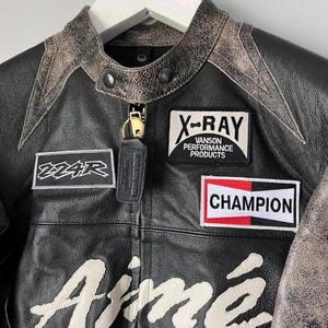 May include: Black leather jacket with distressed accents and patches. Features the text "Aim&eacute;" in white script, along with "X-RAY Vanson Performance Products" and "Champion" patches. Includes a small leather keychain.