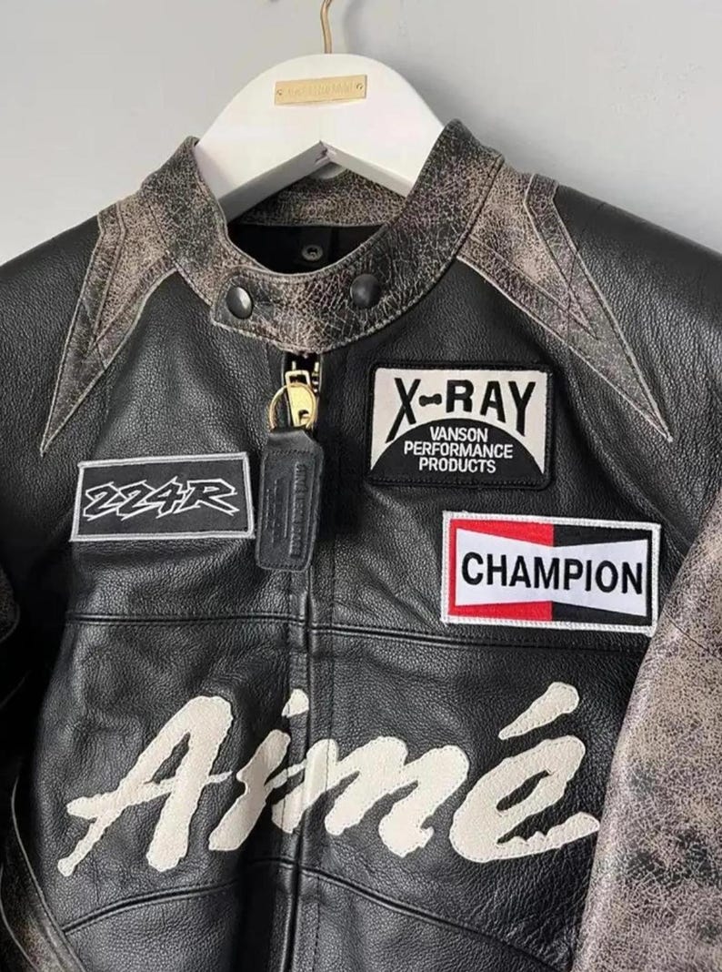 May include: Black leather jacket with distressed accents and patches. Features the text "Aim&eacute;" in white script, along with "X-RAY Vanson Performance Products" and "Champion" patches. Includes a small leather keychain.
