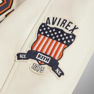 May include: An off-white leather jacket featuring an embroidered patch on the sleeve. The patch displays the text AVIREX, KITH, N.Y., and 1975, alongside a shield graphic with a blue field of white stars and orange and white vertical stripes.
