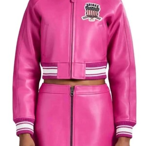 Puede incluir: Conjunto de chaqueta y falda de cuero rosa brillante. La chaqueta tiene cuello, cierre de cremallera y un parche en el pecho. Los pu&ntilde;os y el dobladillo de la chaqueta y la falda tienen detalles a rayas blancas y rosas. La falda tiene cremallera frontal.