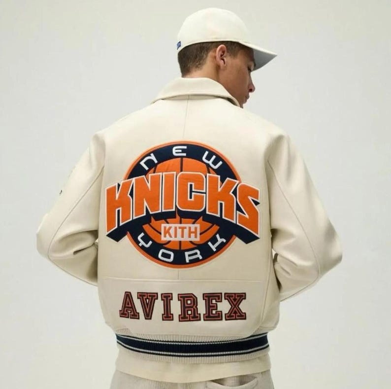 May include: An off-white varsity jacket featuring a large circular graphic on the back with the text NEW YORK KNICKS KITH in orange and navy blue. Below the graphic, the word AVIREX is embroidered in dark red with a navy blue outline.