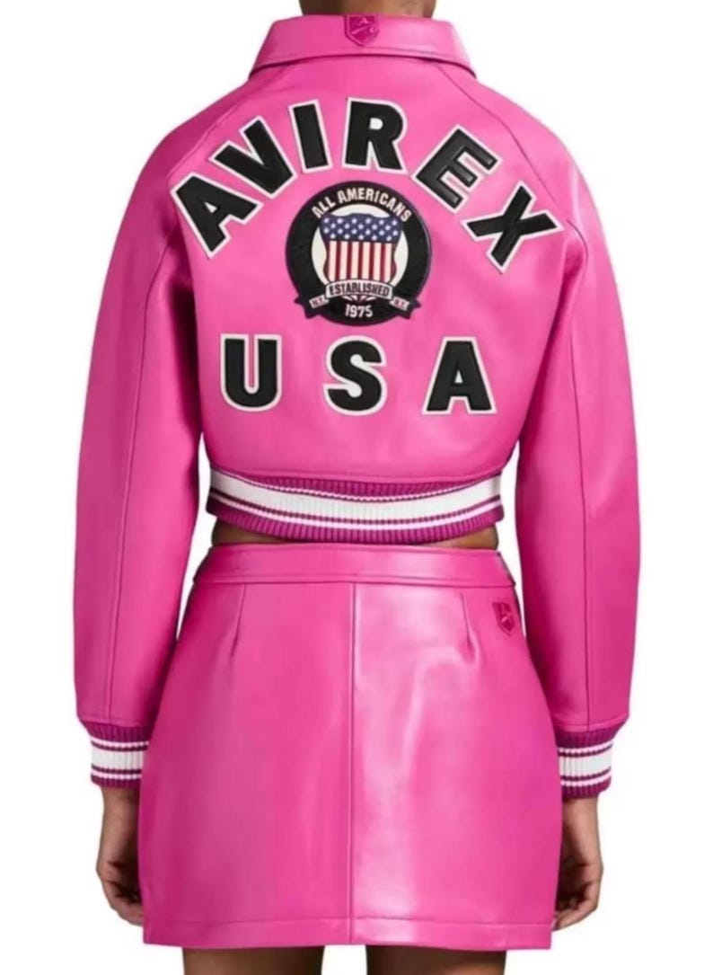 Puede incluir: Un conjunto de chaqueta y falda de cuero rosa brillante. La chaqueta presenta la palabra "AVIREX" en la espalda, junto con un parche que dice "ALL AMERICANS ESTABLISHED 1975 USA". La chaqueta tiene pu&ntilde;os a rayas blancas y negras.