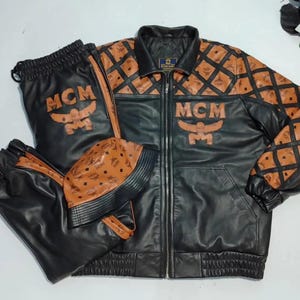 May include: Black leather jacket, pants, and hat set with brown diamond-patterned accents. The jacket and pants feature the "MCM" logo in a gold color. The set includes a zip-up jacket, drawstring pants, and a beanie.