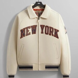 May include: A cream-colored zip-up jacket featuring the text NEW YORK in navy blue and red lettering across the chest. The jacket includes a collared neckline, two front pockets, and navy blue and cream striped ribbed cuffs and hemline.
