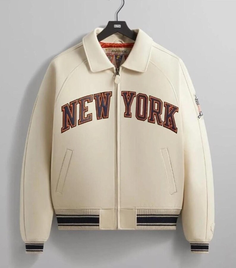 May include: A cream-colored zip-up jacket featuring the text NEW YORK in navy blue and red lettering across the chest. The jacket includes a collared neckline, two front pockets, and navy blue and cream striped ribbed cuffs and hemline.