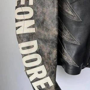 May include: A distressed leather jacket with the words "LEON DORE" in white lettering on the sleeve. The jacket features a black body with white accents and a unique design.