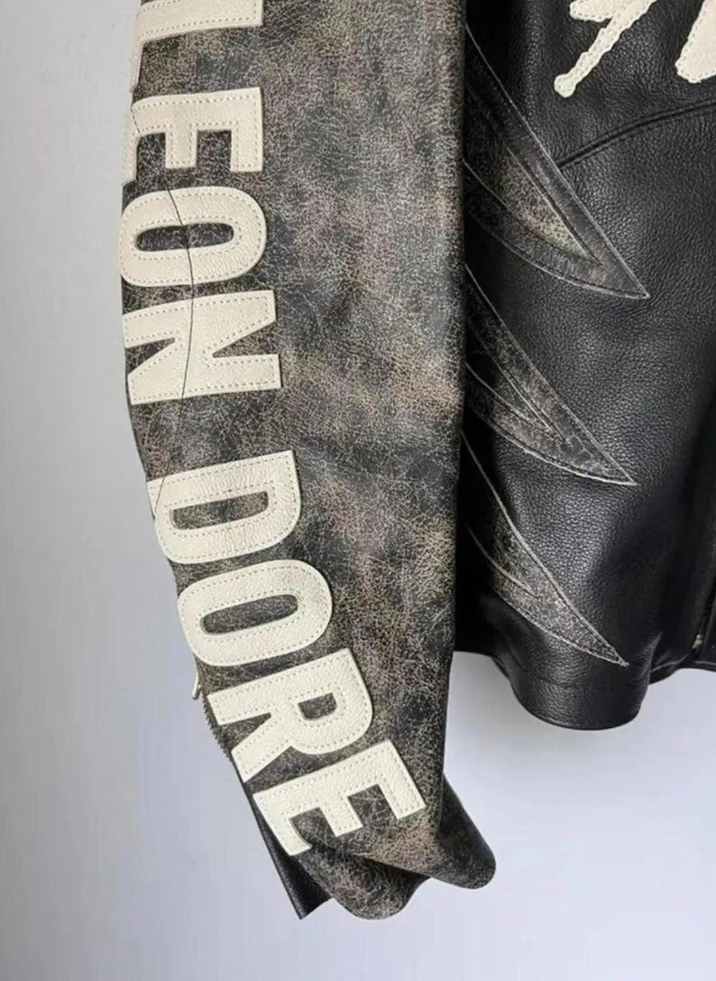 May include: A distressed leather jacket with the words "LEON DORE" in white lettering on the sleeve. The jacket features a black body with white accents and a unique design.