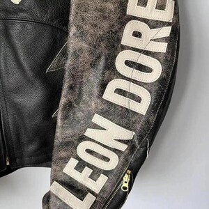 May include: Black leather jacket with the text "LEON DORE" in white block letters on the sleeve. The jacket has a distressed, vintage look with a gold zipper and a star-shaped design.