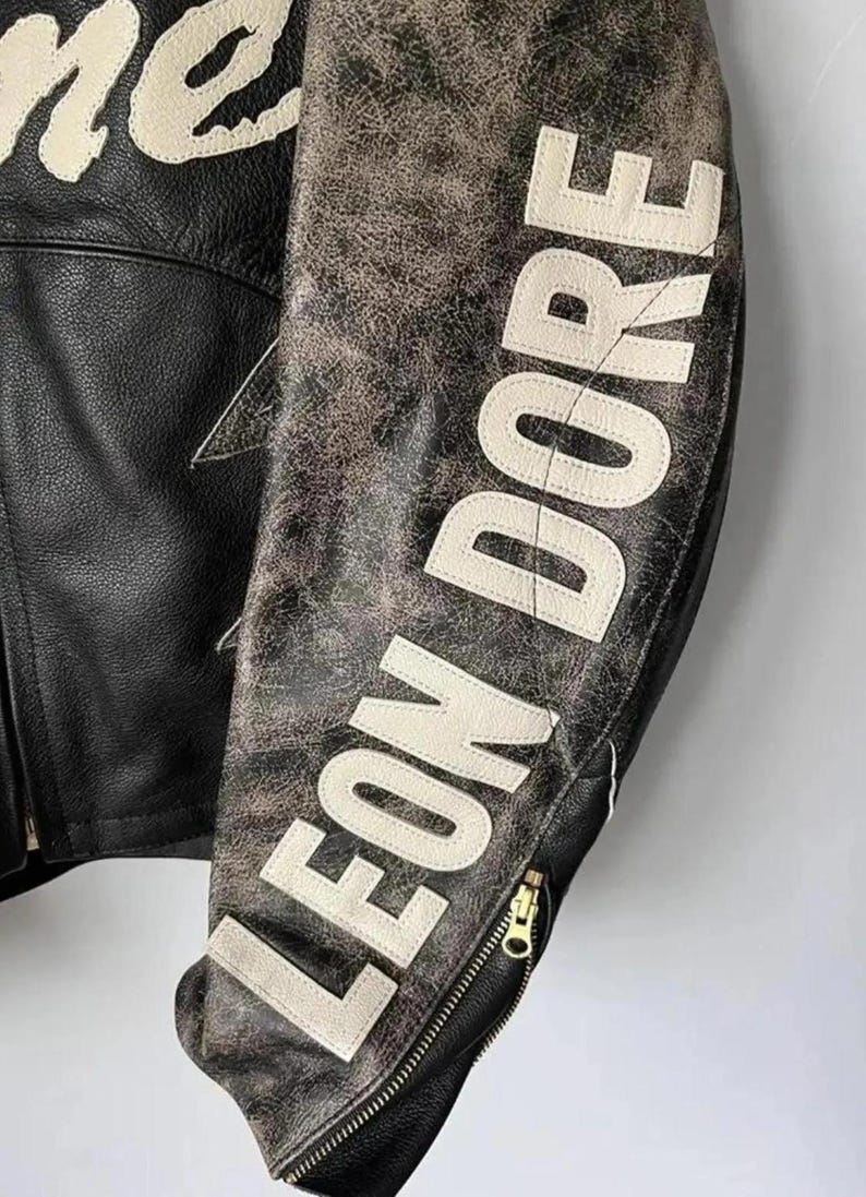 May include: Black leather jacket with the text "LEON DORE" in white block letters on the sleeve. The jacket has a distressed, vintage look with a gold zipper and a star-shaped design.