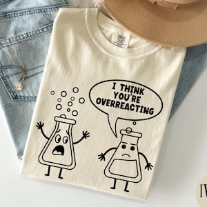 Puede incluir: Camiseta de color marfil con un gráfico negro de dos vasos de dibujos animados. Un vaso reacciona con burbujas, y el otro dice, en un bocadillo, "Creo que estás reaccionando exageradamente". La camiseta está sobre unos vaqueros.