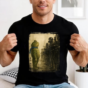 Op de afbeelding: Zwart T-shirt met een grillige illustratie. De afbeelding toont een kikker met een blauwe sjaal die tegenover een rij soldaten met gasmaskers staat. De kikker is groen met bruine vlekken.