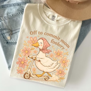 以下が含まれることがあります： スクーターに乗ったアヒルの漫画が描かれたクリーム色のTシャツ。周りには花が描かれ、アヒルの上には「Off to commit mom foolery」の文字。明るい青色のジーンズとベージュの帽子と組み合わせています。