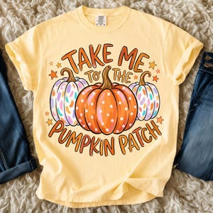 Pode incluir: Uma t-shirt amarelo-claro com um gráfico de três abóboras e o texto "TAKE ME TO THE PUMPKIN PATCH". A abóbora central é laranja com bolinhas brancas, enquanto as duas abóboras laterais apresentam padrões de pinceladas coloridas.