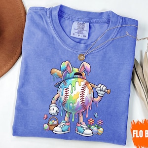 Puede incluir: Una camiseta azul Comfort Colors con un personaje de béisbol con orejas de conejo, sosteniendo un bate. La pelota de béisbol está decorada con colores del arcoíris y pintura goteando. La camiseta tiene una etiqueta pequeña y un collar dorado. El texto "FLO BLUE" está en la parte inferior derecha.