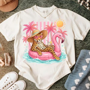 Könnte beinhalten: Weißes T-Shirt mit einem Cartoon-Leoparden, der eine Sonnenbrille und einen Strohhut trägt und auf einem rosa Flamingo-Schwimmer im Wasser liegt. Das Design umfasst Palmen, eine Sonne und ein Getränk. Das Shirt hat einen V-Ausschnitt.