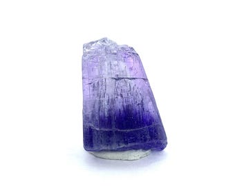 May include: A raw, uncut tanzanite gemstone. The crystal displays a gradient of colors, transitioning from a deep indigo at the base to a lighter lavender at the top. The gemstone is set on a small, white base, with a white background.